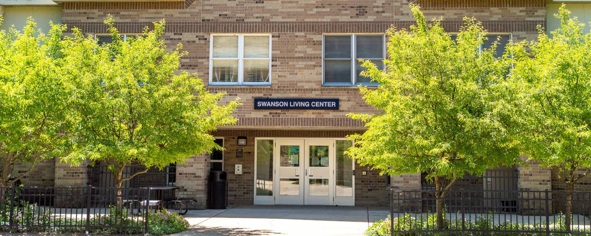 Bright sunny day at Swanson Living Center, showcasing the brick facade, central entrance, and lush green trees enhancing the welcoming atmosphere.